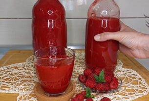 Dve flaše sirupa od malina na stolu kao i čaša i posute maline pored. osoba drži flašu, pozadina je drveni sto i milje na njemu