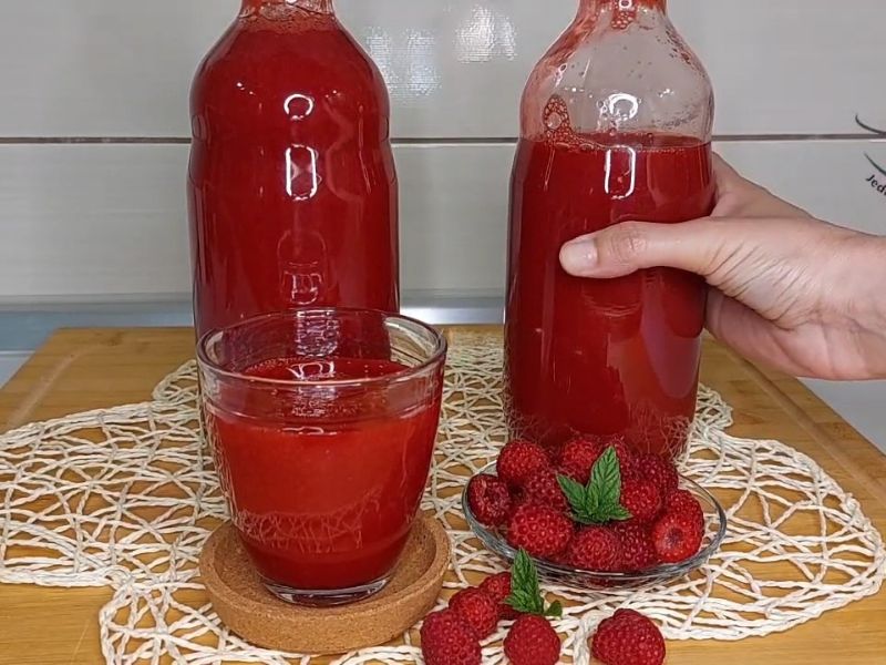 Dve flaše sirupa od malina na stolu kao i čaša i posute maline pored. osoba drži flašu, pozadina je drveni sto i milje na njemu